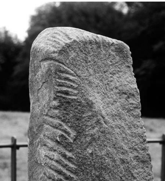 Grobe, verwitterte Steinsäule mit eingeritzten Rillen, unscharfer Hintergrund mit Bäumen und Zaun.